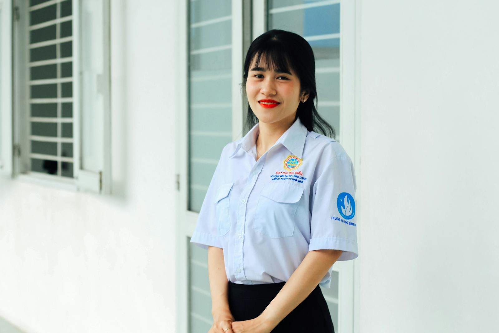 Asian woman in uniform smiling indoors near windows, displaying confidence and warmth.